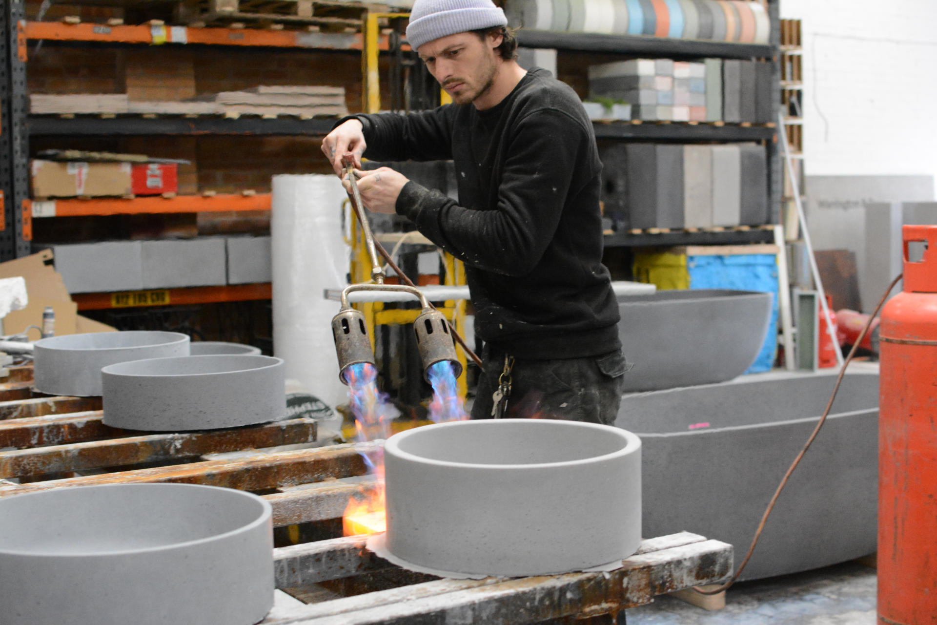 A W+R maker in the Nottingham workshop, flame-finishing a round concrete basin with a twin-headed gas torch, other basins on pallets behind him
